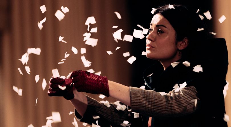 A woman wearing red gloves, pretending to be driving an imaginary car. Paper snow is falling around her.